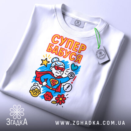 Футболка супер бабуся подарунок, біла, складена на столі, фон світло-сірий, принт з супер бабусею в червоному костюмі, з зеленим ярликом, яскраві кольори принта.