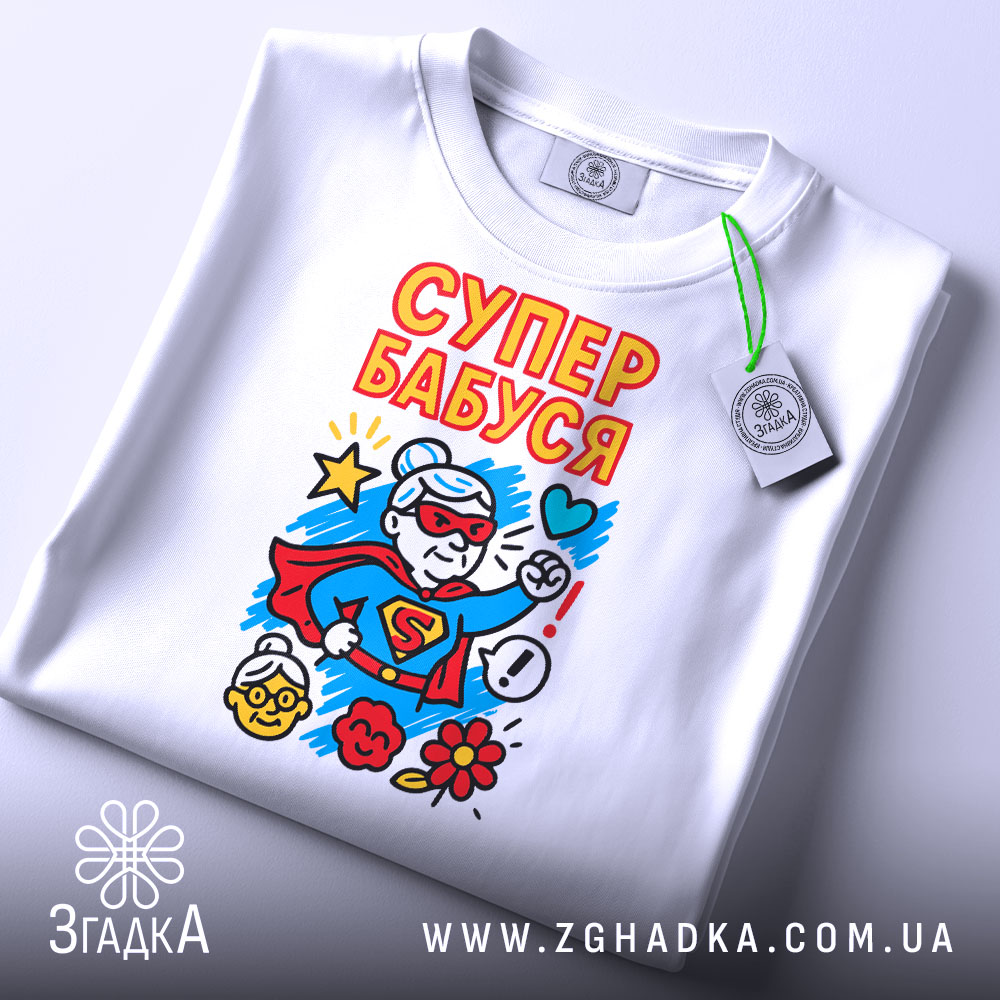 Футболка супер бабуся подарунок, біла, складена на столі, фон світло-сірий, принт з супер бабусею в червоному костюмі, з зеленим ярликом, яскраві кольори принта.