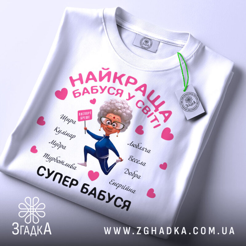 Футболка супер бабуся, біла, складена на столі, фон світло-сірий, принт з бабусею в синьому костюмі, що тримає табличку, з сердечками та словами, що описують її якості, на етикетці видно логотип.