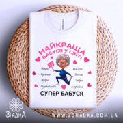 Футболка супер бабуся, біла, складена на плетеній основі, фон світло-сірий, принт з бабусею в синьому костюмі, що тримає табличку, з сердечками та словами, що описують її якості.