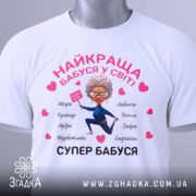 Футболка супер бабуся, біла, складена на столі, фон світло-сірий, принт з бабусею в синьому костюмі, що тримає табличку, з сердечками та словами, що описують її якості, на етикетці видно логотип.