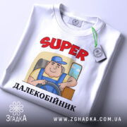 Футболка супер далекобійник білого кольору, зображена з верхнього ракурсу, фон світлий, на футболці принт з мультяшним водієм, текст 'SUPER ДАЛЕКОБІЙНИК' червоного кольору, на футболці є етикетка з зеленим шнурком.