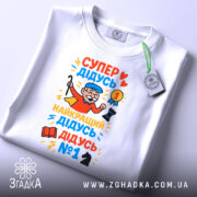 Футболка супер дідусь герой, біла, зображена зверху, на фоні світло-сірого кольору, принт яскравий, з дідусям у червоному плащі, який тримає медаль та шахову фігуру, з тегом на зеленій стрічці.