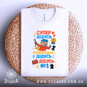 Футболка супер дідусь герой, біла, зображена зверху на плетеній основі, фон світлий, принт яскравий, з дідусям у червоному плащі, який тримає медаль та шахову фігуру.