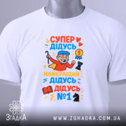 Футболка супер дідусь герой, біла, зображена зверху, на фоні світло-сірого кольору, принт яскравий, з дідусям у червоному плащі, який тримає медаль та шахову фігуру, з тегом на зеленій стрічці.