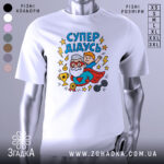 Футболка супер дідусь білого кольору, зображена з фронтального ракурсу на манекені, фон світло-сірий, принт з веселим дідусем у червоному плащі та хлопчиком, що тримається за його плечі, оточений зірками та блискавками.