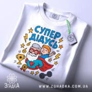 Футболка супер дідусь білого кольору, зображена зверху, на ній видно принт з дідусем та хлопчиком, а також етикетка з зеленим шнурком, фон світло-сірий, деталі принта яскраві та кольорові.