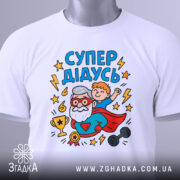 Футболка супер дідусь білого кольору, зображена зверху, на ній видно принт з дідусем та хлопчиком, а також етикетка з зеленим шнурком, фон світло-сірий, деталі принта яскраві та кольорові.
