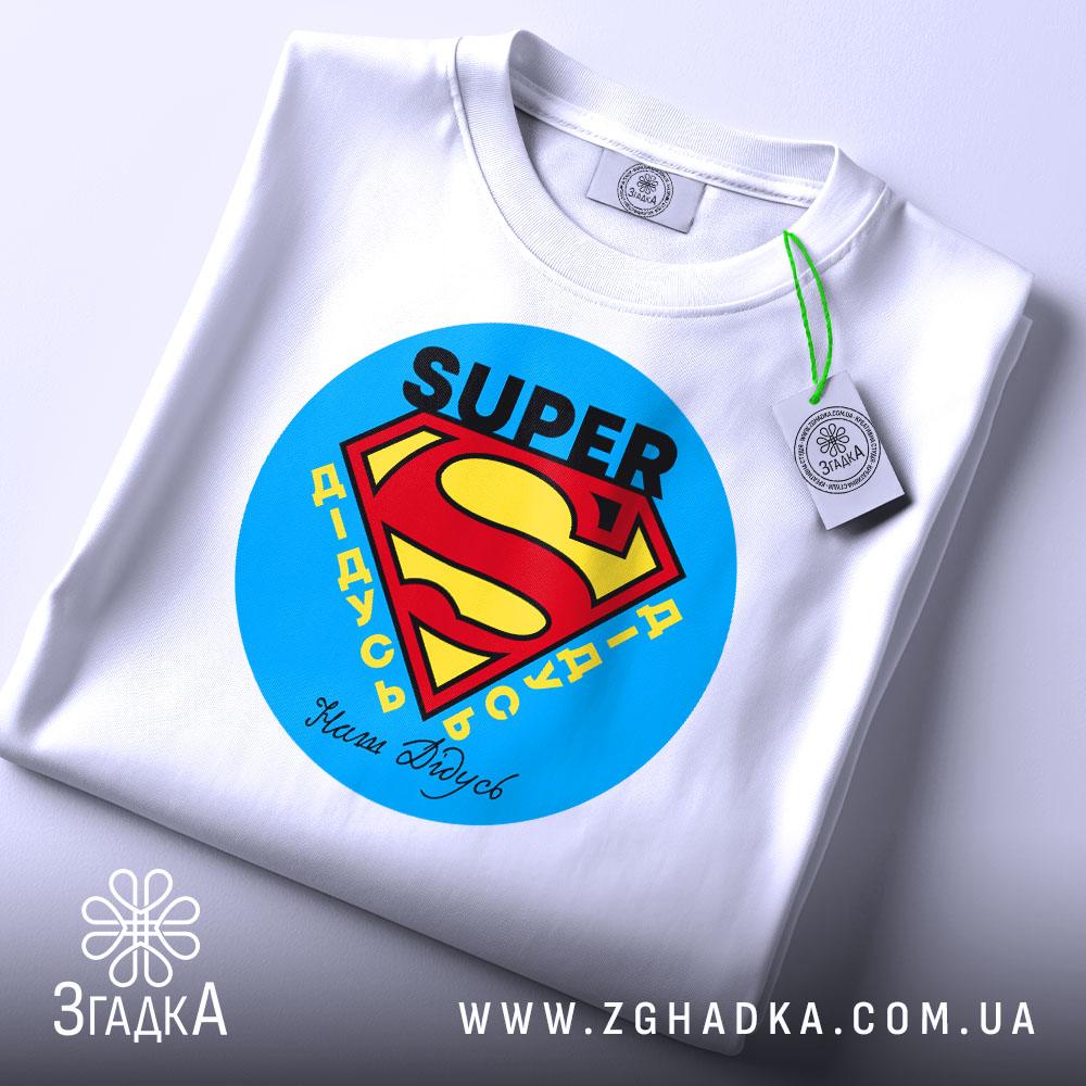 Футболка супер дідусь, біла, складена на столі, фон світло-сірий, принт з логотипом супермена, текст 'SUPER' та 'Наші дідусі' під ним, з етикеткою на рукаві.