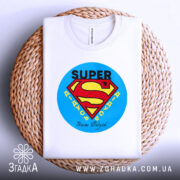 Футболка супер дідусь, біла, складена на плетеній кошику, фон світло-сірий, принт з логотипом супермена, текст 'SUPER' та 'Наші дідусі' під ним.