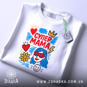 Футболка супер мама, білого кольору, складена на плоскій поверхні, фон світло-сірий, принт яскравий з червоним, жовтим, блакитним кольорами, зображує жінку з блакитним волоссям у червоних окулярах, серце, квітка та блискавка, на комірці є етикетка.