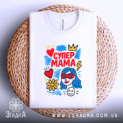 Футболка супер мама, білого кольору, складена на круглому плетеному кошику, фон білий, принт яскравий з червоним, жовтим, блакитним кольорами, зображує жінку з блакитним волоссям у червоних окулярах, серце, квітка та блискавка.