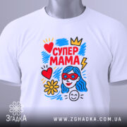 Футболка супер мама, білого кольору, складена на плоскій поверхні, фон світло-сірий, принт яскравий з червоним, жовтим, блакитним кольорами, зображує жінку з блакитним волоссям у червоних окулярах, серце, квітка та блискавка, на комірці є етикетка.