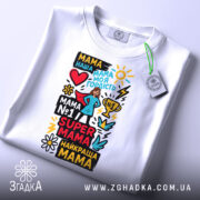 Футболка супер мама, біла, складена на столі, фон світло-сірий, принт кольоровий з текстом та малюнками, прикріплена етикетка зеленого кольору.