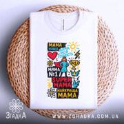 Футболка супер мама, біла, складена на плетеній кошику, фон світло-сірий, принт кольоровий з текстом та малюнками, включаючи серце, зірки та квіти.