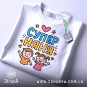 Футболка супер няня, біла, зображена на столі, ракурс зверху, фон світло-сірий, принт з двома дітьми, які радісно піднімають руки, текст 'СУПЕР НЯНЯ' у яскравих кольорах, з етикеткою.