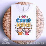 Футболка супер няня, біла, зображена на плетеному кошику, фон світло-сірий, принт з двома дітьми, які радісно піднімають руки, текст 'СУПЕР НЯНЯ' у яскравих кольорах.