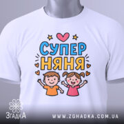 Футболка супер няня, біла, зображена на столі, ракурс зверху, фон світло-сірий, принт з двома дітьми, які радісно піднімають руки, текст 'СУПЕР НЯНЯ' у яскравих кольорах, з етикеткою.