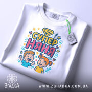 Футболка супер няня, білого кольору, складена на поверхні, фон світло-сірий, принт з дітьми та написом 'СУПЕР НЯНЯ', з етикеткою на рукаві.