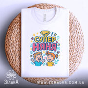 Футболка супер няня, білого кольору, складена на плетеній кошику, фон світло-сірий, принт з дітьми та написом 'СУПЕР НЯНЯ' у яскравих кольорах.
