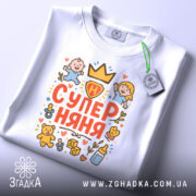 Футболка супер няня білого кольору, складена на поверхні, з видимим етикеткою, принт з малюнками дітей та іграшок, розташований по центру.