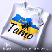 Футболка супер тато білого кольору, складена на поверхні, фон світло-сірий, принт з синім та жовтим кольорами, силует супергероя та напис 'Супер Тато' з елементами декору.