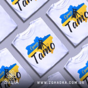 Футболка супер тато білого кольору, зображена на манекені, фон світло-сірий, принт включає синій та жовтий кольори з силуетом супергероя та написом 'Супер Тато'.