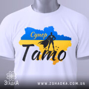 Футболка супер тато білого кольору, складена на поверхні, фон світло-сірий, принт з синім та жовтим кольорами, силует супергероя та напис 'Супер Тато' з елементами декору.