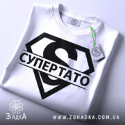 Футболка супер тато супермен, біла, складена на плоскій поверхні, фон світло-сірий, принт чорний з великим літерою 'S' та написом 'СУПЕРТАТО', на комірці є етикетка з зеленим шнурком.