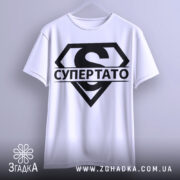 Футболка супер тато супермен, біла, підвішена на вішалці, фон світло-сірий, принт чорний з великим літерою 'S' та написом 'СУПЕРТАТО'.