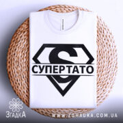 Футболка супер тато супермен, біла, складена на плоскій поверхні, фон світло-сірий, принт чорний з великим літерою 'S' та написом 'СУПЕРТАТО', розташована на плетеній основі.