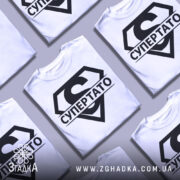 Футболка супер тато супермен, біла, зображена з близького ракурсу, на манекені, фон світло-сірий, принт чорний з великим літерою 'S' та написом 'СУПЕРТАТО'.