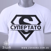 Футболка супер тато супермен, біла, складена на плоскій поверхні, фон світло-сірий, принт чорний з великим літерою 'S' та написом 'СУПЕРТАТО', на комірці є етикетка з зеленим шнурком.