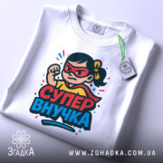 Футболка супер внучка білого кольору, складена на столі, з принтом дівчинки-супергероя, на якій видно червоні окуляри та жовту футболку, на етикетці зелена стрічка, фон світлий.