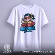 Футболка супер внучка білого кольору, підвішена на вішалці, з принтом дівчинки-супергероя, фон світлий, принт яскравий, видно деталі принта.