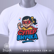 Футболка супер внучка білого кольору, складена на столі, з принтом дівчинки-супергероя, на якій видно червоні окуляри та жовту футболку, на етикетці зелена стрічка, фон світлий.