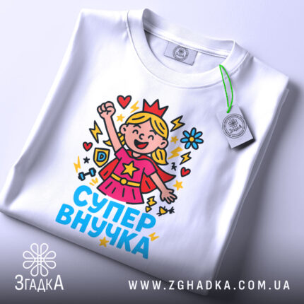 Футболка супер внучка білого кольору, складена на поверхні, фон світло-сірий, принт з дівчинкою-супергероєм, що піднімає руку, з яскравими кольорами та написом 'СУПЕР ВНУЧКА', на комірці є етикетка.