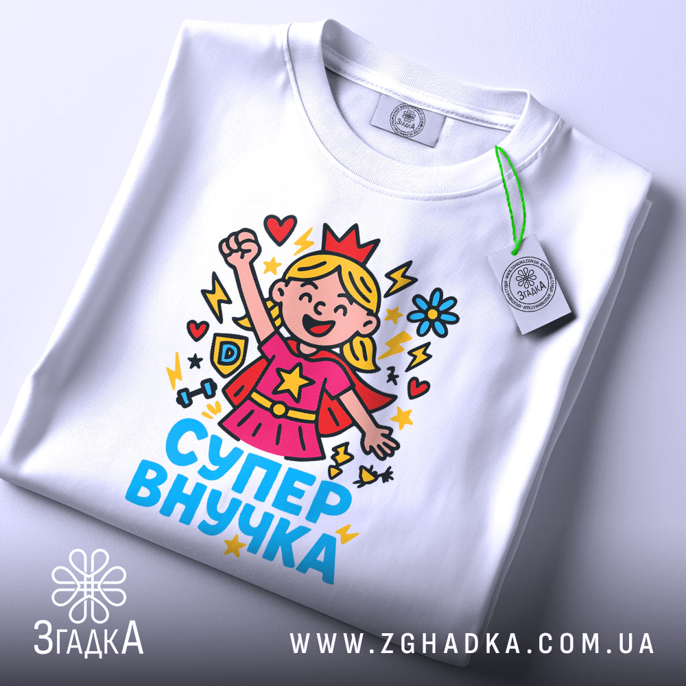 Футболка супер внучка білого кольору, складена на поверхні, фон світло-сірий, принт з дівчинкою-супергероєм, що піднімає руку, з яскравими кольорами та написом 'СУПЕР ВНУЧКА', на комірці є етикетка.