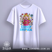 Футболка супер внучка білого кольору, підвішена на вішалці, фон світло-сірий, принт з дівчинкою-супергероєм, що піднімає руку, з яскравими кольорами та написом 'СУПЕР ВНУЧКА'.