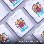 Футболка супер внучка білого кольору, зображена на манекені з правого боку, фон світло-сірий, принт з веселою дівчинкою-супергероєм, що піднімає руку, з яскравими кольорами та написом 'СУПЕР ВНУЧКА'.