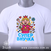 Футболка супер внучка білого кольору, складена на поверхні, фон світло-сірий, принт з дівчинкою-супергероєм, що піднімає руку, з яскравими кольорами та написом 'СУПЕР ВНУЧКА', на комірці є етикетка.