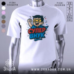 Футболка супер внук білого кольору, зображена на манекені, ракурс спереду, фон світло-сірий, принт з веселим персонажем у масці та плащі, з піднятою рукою, текст 'СУПЕР ВНУК' яскравими кольорами.