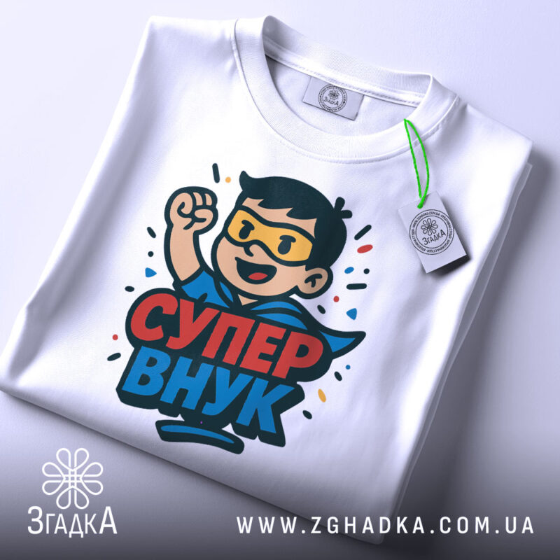 Футболка супер внук білого кольору, зображена на столі, ракурс зверху, фон світло-сірий, принт з персонажем у масці та плащі, текст 'СУПЕР ВНУК', на футболці є етикетка з зеленим шнурком.