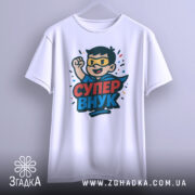 Футболка супер внук білого кольору, зображена на вішалці, ракурс спереду, фон світло-сірий, принт з персонажем у масці та плащі, текст 'СУПЕР ВНУК' яскравими кольорами.