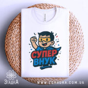 Футболка супер внук білого кольору, зображена на плетеній кошику, ракурс зверху, фон світло-сірий, принт з персонажем у масці та плащі, текст 'СУПЕР ВНУК' яскравими кольорами.