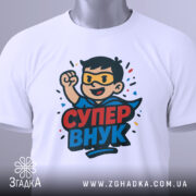 Футболка супер внук білого кольору, зображена на столі, ракурс зверху, фон світло-сірий, принт з персонажем у масці та плащі, текст 'СУПЕР ВНУК', на футболці є етикетка з зеленим шнурком.