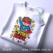 Футболка супер внук білого кольору, складена на поверхні, фон світло-сірий, принт з супергероєм у червоному плащі, що піднімає кулак, з трофеєм та блискавками.