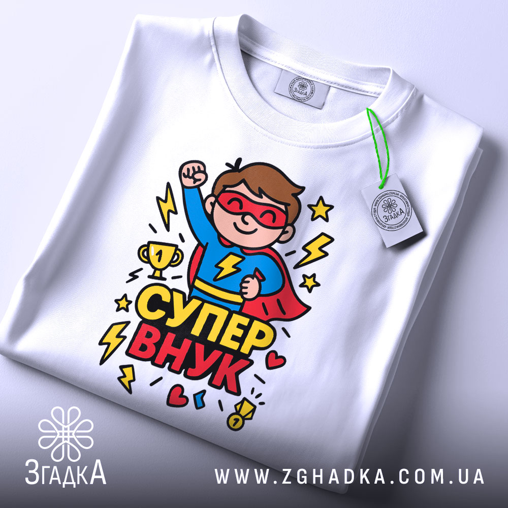 Футболка супер внук білого кольору, складена на поверхні, фон світло-сірий, принт з супергероєм у червоному плащі, що піднімає кулак, з трофеєм та блискавками.