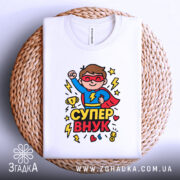 Футболка супер внук білого кольору, складена на плетеній кошику, фон світло-сірий, принт з супергероєм у червоному плащі, що піднімає кулак, з трофеєм та блискавками.