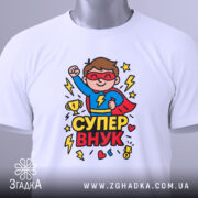 Футболка супер внук білого кольору, складена на поверхні, фон світло-сірий, принт з супергероєм у червоному плащі, що піднімає кулак, з трофеєм та блискавками.